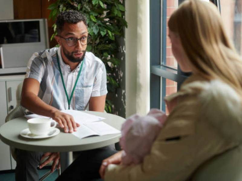 A young gentleman sits across a table from a young woman with a baby in her lap. They are discussing papers on a table.