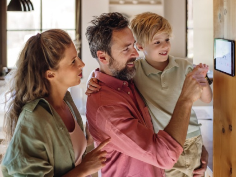 A father holding his son up, showing him a smart thermostat on the wall, while the mother looks on.