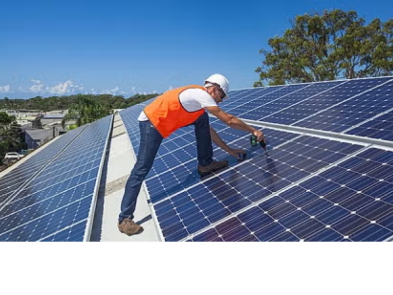 A worker in an orange vest and white hard hat drills into a roof top solar panel.