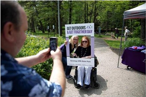 A man is taking a photo of two women, one of the women is in a wheelchair.  The women are framing themselves with a sign that says Get Outdoors and Get Together