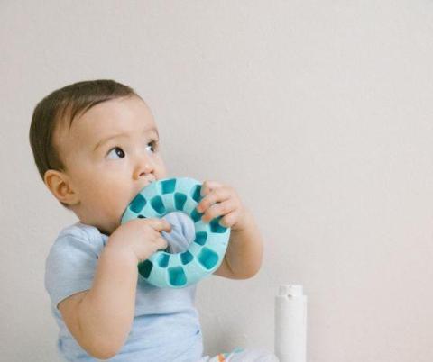 Infant sits up cross-legged and uses both hands to put plush toy ring in mouth.